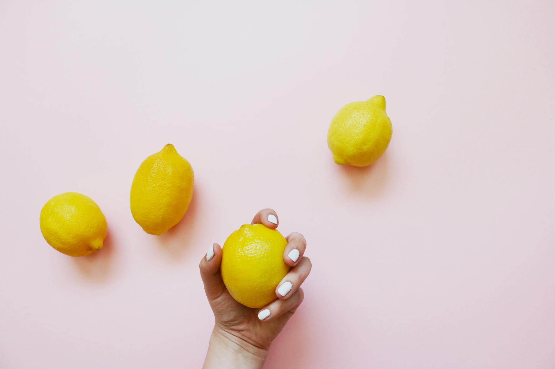 A hand holding a fresh lemon on a soft pink background surrounded by additional lemons, symbolizing the simplicity and refreshing nature of solo pleasure